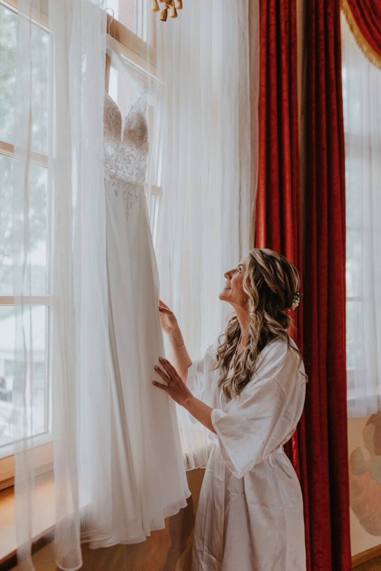 Braut betrachtet ihr Brautkleid am Fenster